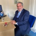 A man smiling as he's sat down on a chair next to a desk