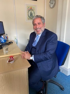 A man smiling as he's sat down on a chair next to a desk