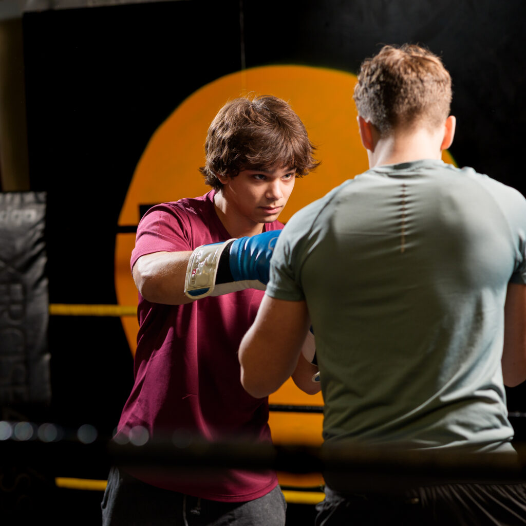 Two young boxers spar inside a ring, one in a maroon shirt wearing blue gloves facing a partner with his back to camera.