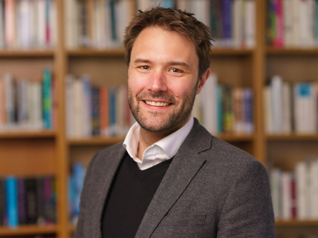 Smiling man in a gray blazer stands in a library with blurred bookshelves behind him.