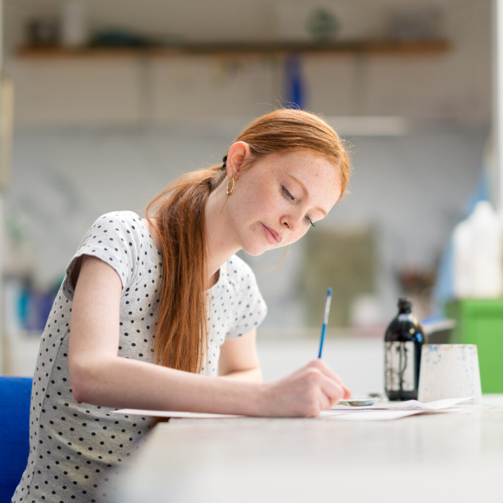 Woman with red hair writing at a white table in a bright room, focusing on her task with a pen and papers in front of her