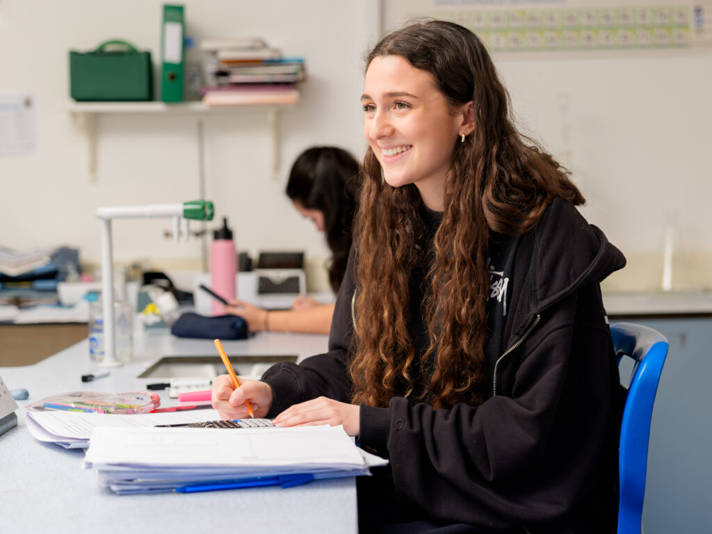 Young woman with long hair sits at a lab-style desk, smiling while writing with an orange pencil at papers and notebooks in front of her.
