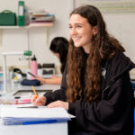 Young woman with long hair sits at a lab-style desk, smiling while writing with an orange pencil at papers and notebooks in front of her.