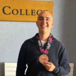 Smiling young man indoors, proudly holding a bronze medal on a pink-and-black ribbon in front of a college sign.