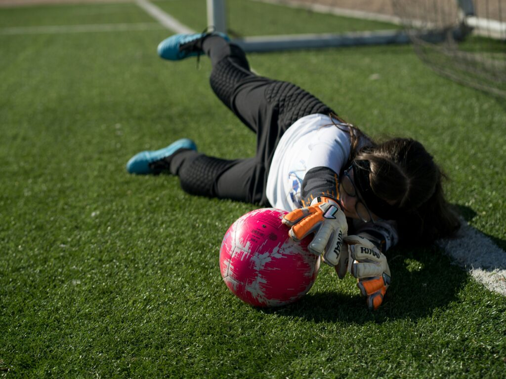 Soccer goalkeeper dives on artificial turf to stop a shot, with a pink ball and gloves in the foreground.
