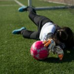 Soccer goalkeeper dives on artificial turf to stop a shot, with a pink ball and gloves in the foreground.