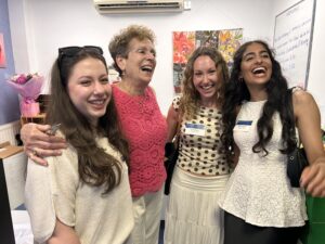 Four women smile and pose together indoors, with a pink bouquet on the left and a whiteboard in the background, all wearing dresses.