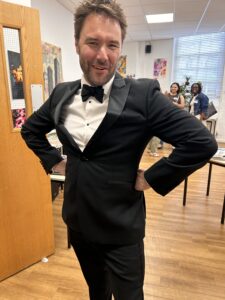 Man in a black tuxedo with bow tie posing with hands on hips, smiling in a room with artwork and other people in the background.