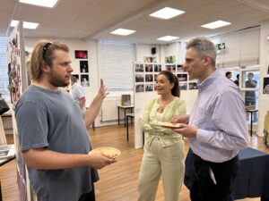 Man with long hair and beard gestures while talking to a man in a blue checkered shirt and a smiling woman at an art exhibit.