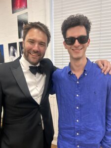 Two men posing for a photo indoors; one in a black tuxedo with a bow tie, the other in a blue shirt wearing sunglasses and smiling.