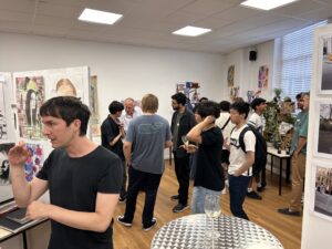 Group of people at an indoor art exhibition, viewing colorful wall artworks with a metal table and wine glass in the foreground.