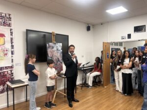 Man in a black suit speaks into a microphone at the front of a classroom while a group of students listens nearby, artworks on the wall and a large screen behind him.