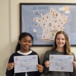 Two girls stand side by side, smiling, holding certificates in front of a framed France map mural in a classroom.