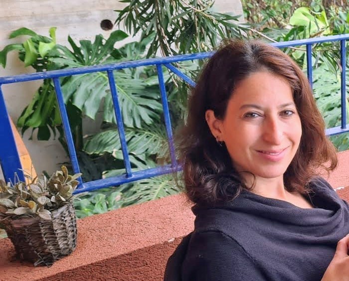 Smiling woman with shoulder-length brown hair sits on a terracotta bench outdoors, with a blue railing and greenery behind.