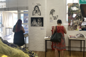 Two women browse artwork in a bright gallery with white brick walls and wall-mounted prints?on display.