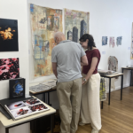 A man and a woman examine mixed-media textile art panels hanging on a gallery wall.