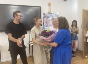 A small celebration: a woman in a beige dress hands a bouquet to a woman in a blue dress while others smile nearby, wine glasses in hand.
