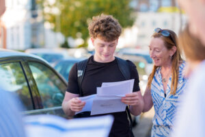 Young man in a black t-shirt holds papers while a woman with sunglasses on her head smiles beside a parked car on a sunny street.