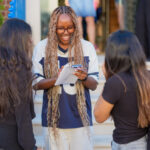 A smiling student with long braided hair holds papers and a phone while talking with peers outside a campus building.