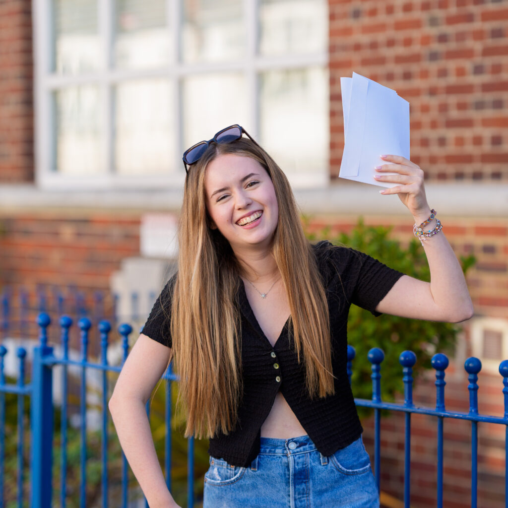Young woman with sunglasses on her head, holding white papers aloft and smiling outside a brick building by a blue fence.