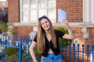 Young woman with sunglasses on her head, holding white papers aloft and smiling outside a brick building by a blue fence.