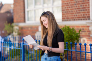 Young woman with long hair reads a sheet of paper outdoors beside a blue fence and brick building.
