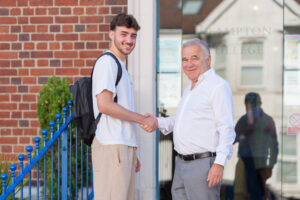 Two men shake hands outside a brick building, one with a backpack and smiles at the camera.