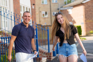 Two young adults standing outdoors, smiling and holding papers, with a blue metal railing and brick buildings behind them.